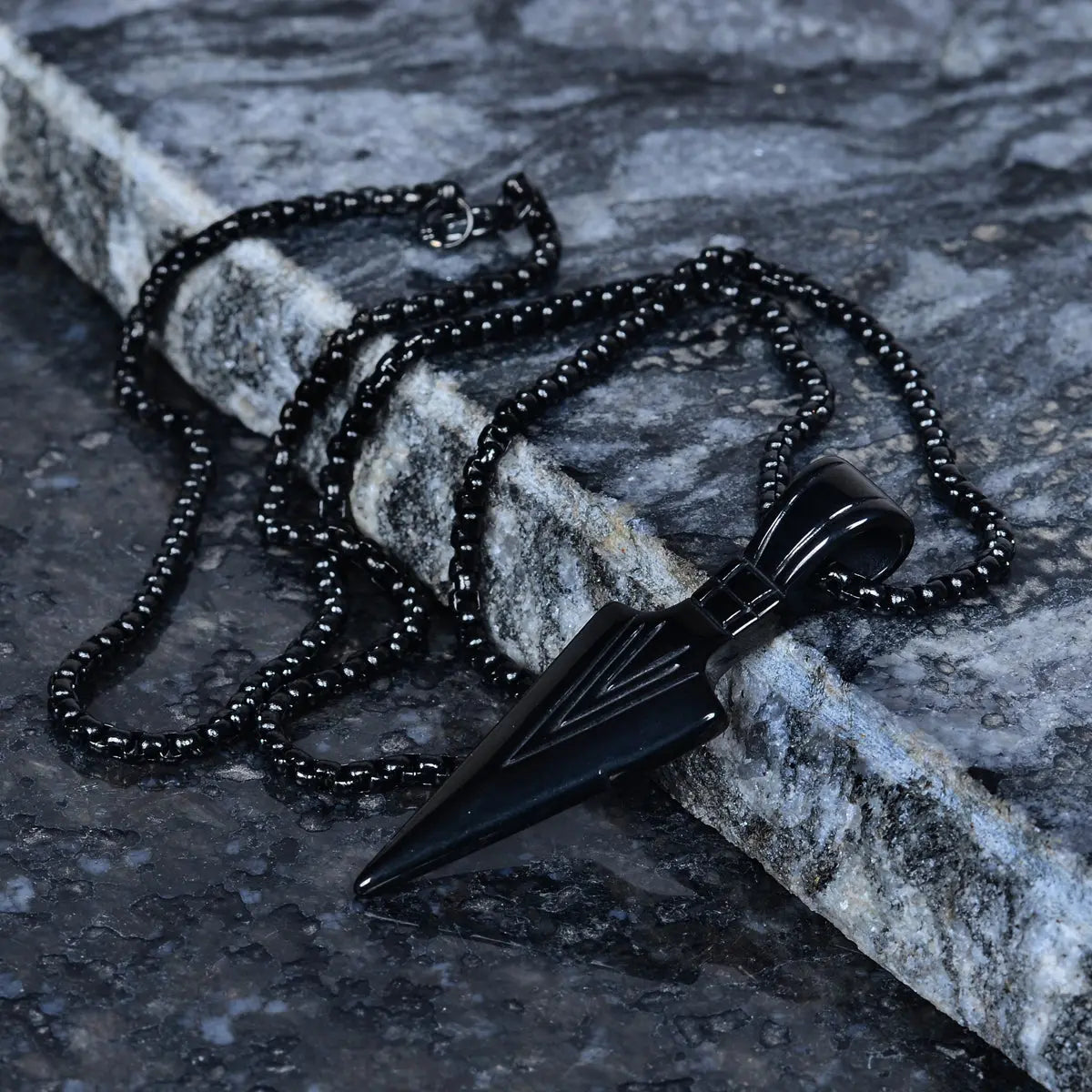 Black dagger-shaped pendant for men on a chain lying on a stone surface by Supreme Stud